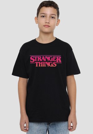 Junge trägt ein schwarzes "Stranger Things"-T-Shirt und hellblaue Jeans, steht vor einem einfarbigen Hintergrund, schaut nach vorne mit neutralem Ausdruck.