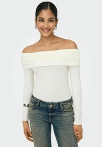 Hvid off-the-shoulder top med samlet tekstur, lange ærmer, parret med blå denim jeans. Bemærkelsesværdig sølvaccent om håndleddet.
