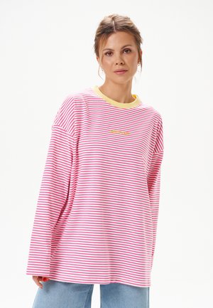 Femme aux cheveux châtain clair portant une chemise à manches longues rose et blanche rayée oversize avec un col jaune, debout sur fond blanc.