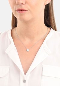 Silverhalsband med en cirkulär hänge som har en klar sten, bärs över en vit skjorta med krage, vilket visar en delikat kedjedesign.