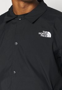 Veste noire North Face avec col et boutons-pression marqués, portée par une personne avec le cou et une partie de la poitrine visibles.