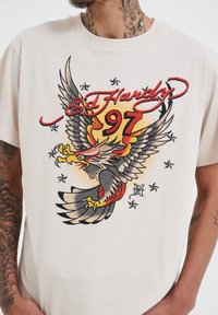 Krémové tričko s multicolorovým grafikou orla, červeným a černým textem s nápisy "Ed Hardy" a "97" a černými hvězdnými akcenty kolem designu.
