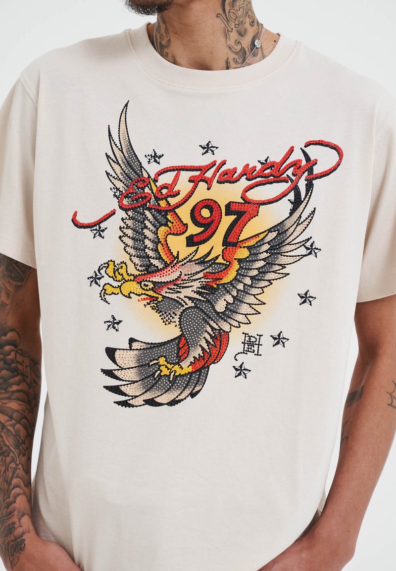 Krémové tričko s multicolorovým grafikou orla, červeným a černým textem s nápisy "Ed Hardy" a "97" a černými hvězdnými akcenty kolem designu.