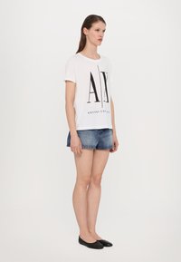 White short-sleeve t-shirt with black "AX" graphic and "Armani Exchange" text, paired with denim shorts and black flats.