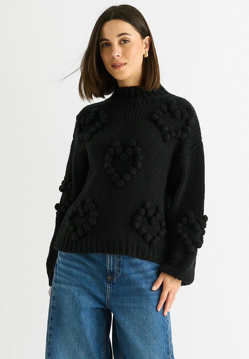 Black knitted sweater with textured puff heart designs, ribbed neckline, and long sleeves. Paired with light blue high-waisted jeans.