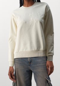 Mujer vestida con un sudadero color crema claro con la palabra "LOVE" en relieve en el pecho y vaqueros cargo azul deslavado, de pie con las manos relajadas a los lados.