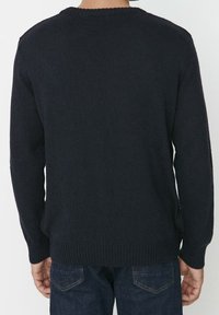 Marinblå stickad tröja med rund halsringning, ribbade ärmslut och nederkant, med en texturerad yta, visad bakifrån. Matchad med mörka jeans.