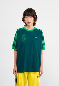Žalias trumpas rankovėmis Adidas marškinėliai su kontrastingais žaliais apykaklės ir rankovių kraštais, puikuojantys Adidas logotipu ir grafikos atspaudais priekyje.