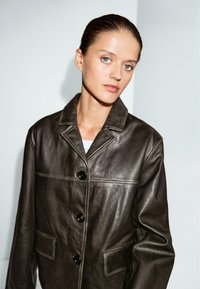 Žena s uhlazenými vlasy nosí tmavě hnědou koženou bundu s velkými knoflíky a stojí před obyčejným světle šedým pozadím.