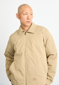 Man with shaved head wearing beige zip-up jacket with collar, standing against a plain white background, hands in pockets, looking left.