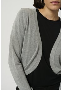 Femme portant un cardigan court gris argenté sur un haut noir avec des boucles d'oreilles anneaux argentés, montrée de l'épaule au menton.