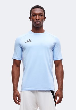Homme debout portant un maillot de sport bleu clair avec un logo noir sur la poitrine et un short de sport blanc avec une bande noire.