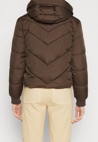 Brown puffer jacket with a hood, featuring a chevron quilted design, elastic cuffs, and a cropped fit. Worn over beige cargo pants.