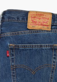 Jean Levi's 505 en denim bleu moyen, doté de coutures jaunes, d'un design classique à cinq poches, et d'un patch en cuir marron avec un détail de logo rouge.