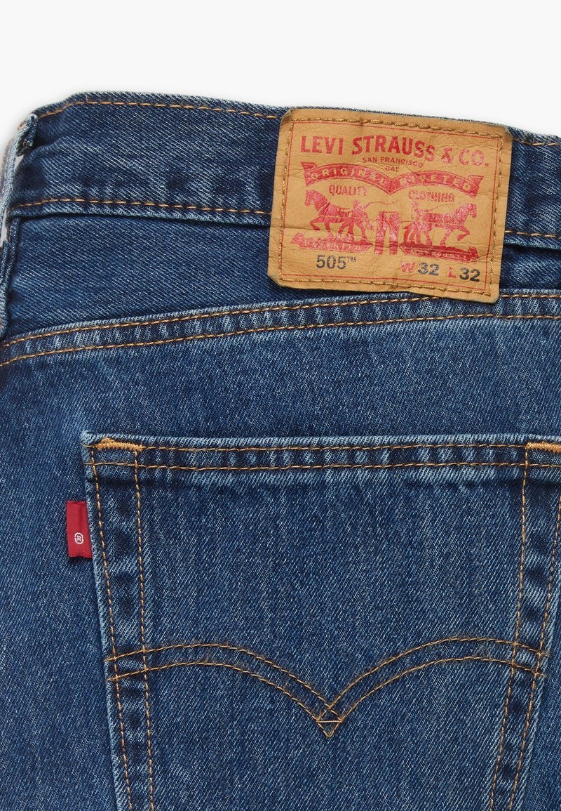 Levi's 505 Jeans aus mittelblauem Denim mit gelber Naht, klassischem Fünf-Taschen-Design und einem beigen Lederpatch mit rotem Logodetail.
