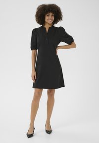 Crna haljina s kratkim nabranim rukavima, ovratnikom na dugmad i ravnim krojem. Tkanina izgleda glatko, uz stilske crne štikle.