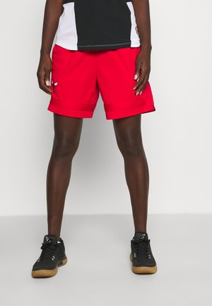 Personne portant un short de sport rouge, un haut de sport noir et blanc, et des baskets noires à semelles beiges, debout devant un fond uni.
