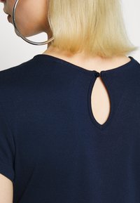 Granatowa bluzka z krótkimi rękawami i wycięciem w kształcie kropli na plecach, z małym zapięciem na guzik. Gładka, elastyczna tekstura materiału.
