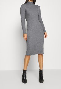 Robe grise à manches longues côtelées avec un col montant, silhouette ajustée et longueur mi-mollet. Associée à des bottines noires.