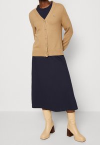 Kamelfarvet cardigan med v-hals og knaplukning, kombineret med en marineblå knælang kjole. Beige støvler med hæl og firkantet tå fuldender outfittet.