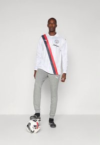 Fehér, hosszú ujjú ing piros és sötétkék átlós csíkokkal, szürke sportnadrág és fekete focicipő; a padlón egy focilabda.