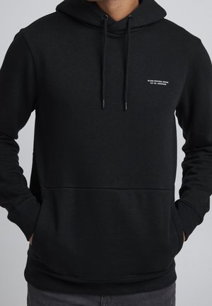 Mann trägt schwarzen Hoodie mit Fronttasche und kleinem weißen Text "Blend Original Denim Est. 93 - Denmark" auf der Brust vor einfachem Hintergrund.