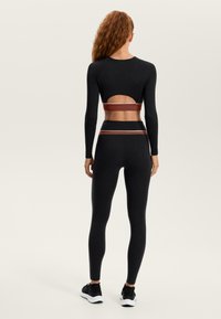 Fekete hosszú ujjú felső nyitott háttal és barna derékpánttal, fekete leggingsszel párosítva. Könnyű, testhez álló kialakítás.