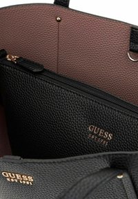 Intérieur d'un sac fourre-tout en cuir texturé noir, comprenant une pochette zippée noire avec logo doré "GUESS EST 1981" et une doublure mauve douce.