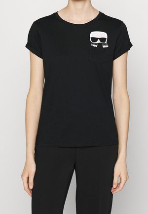 Camiseta de algodón negra con mangas enrolladas, que presenta un diseño gráfico en el bolsillo blanco de un rostro con gafas y manos debajo.
