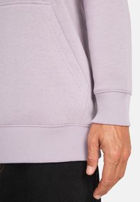 Felpa viola chiaro realizzata in tessuto morbido, con tasca a marsupio, polsini a coste e una texture liscia. Pantaloni neri parzialmente visibili.