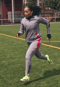 Conjunto atlético en gris con acentos rojos; chaqueta ajustada y leggings, con franjas y logo en negro. Zapatillas brillantes amarillas con detalles en negro.
