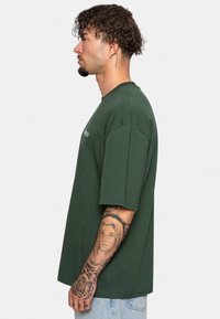Grøn bomulds T-shirt med korte ærmer og rund hals, der har en afslappet pasform. Detaljeret tatovering synlig på underarmen.