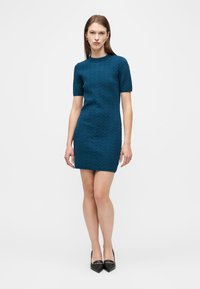 Γυναίκα που φοράει εφαρμοστό, κοντομάνικο, μέχρι το γόνατο φόρεμα σε τυρκουάζ χρώμα με διακριτικό ζιγκ-ζαγκ σχέδιο και μαύρες μυτερές γόβες, στέκεται σε λευκό φόντο.