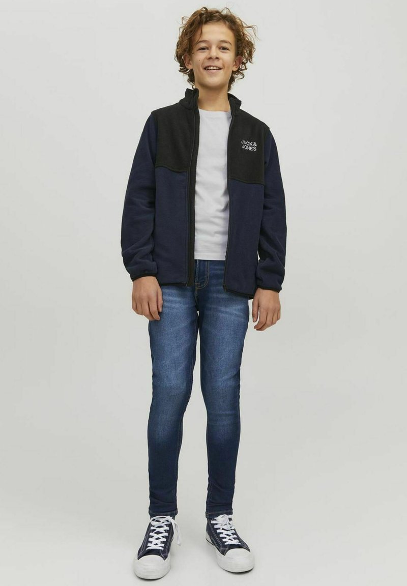 Marinblå och svart zip-up fleecejacka med hög krage, logotyp på bröstet, matchad med ljusblå denimjeans och marinblå sneakers.