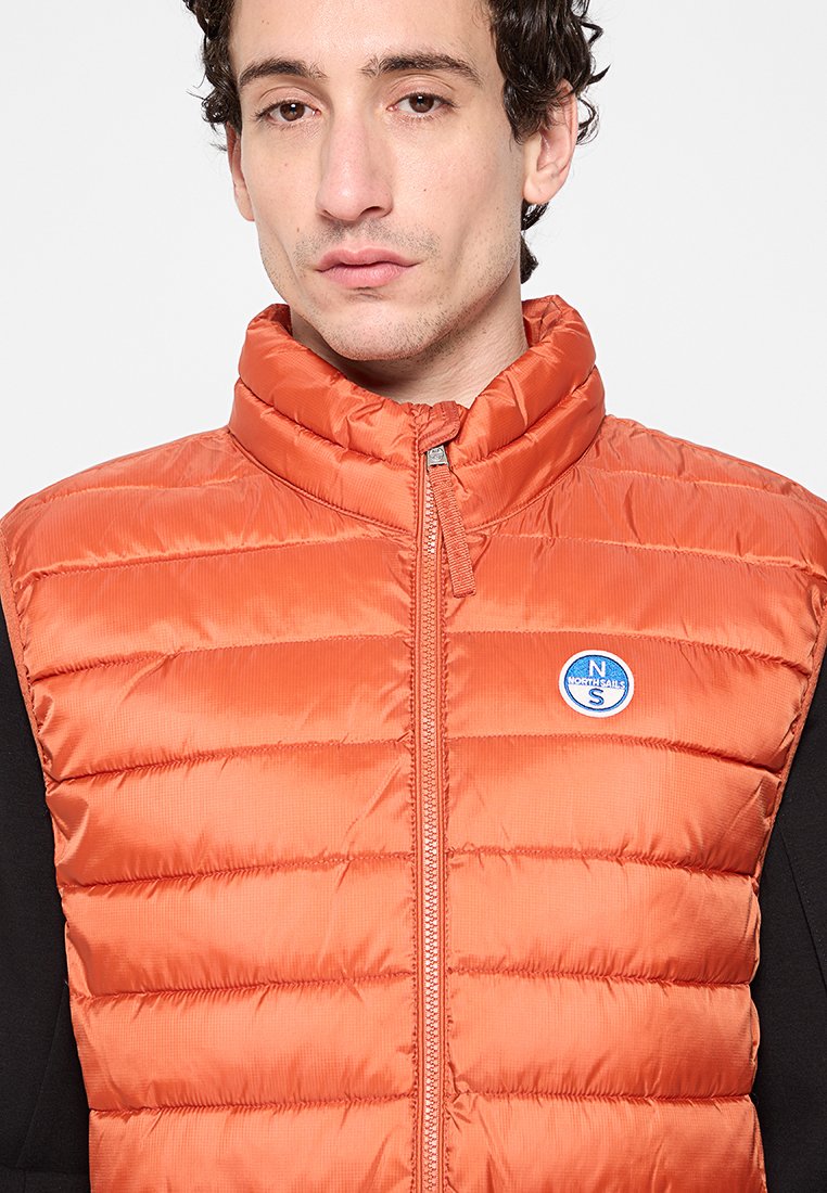 Kurze orange Steppweste mit hohem Kragen, Reißverschluss und einem runden blauen Logo auf der linken Brust. Der Stoff hat eine glatte, glänzende Oberfläche.