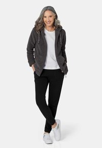 Veste polaire noire avec capuche, chemise grise et pantalon noir. Le modèle porte des baskets blanches. Texture douce avec un design décontracté.