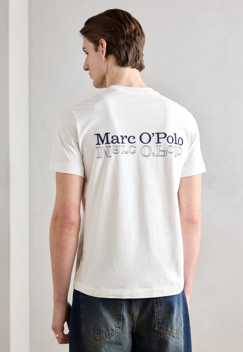 Hvid bomulds T-shirt med et marineblåt logo på ryggen. Korte ærmer og en klassisk pasform, stylet med blå denimjeans.