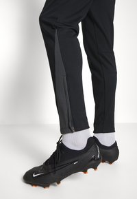 Benenzicht met zwarte sportbroeken met zijrits en zwarte Nike Phantom voetbalschoenen met witte sokken op een witte vloer.