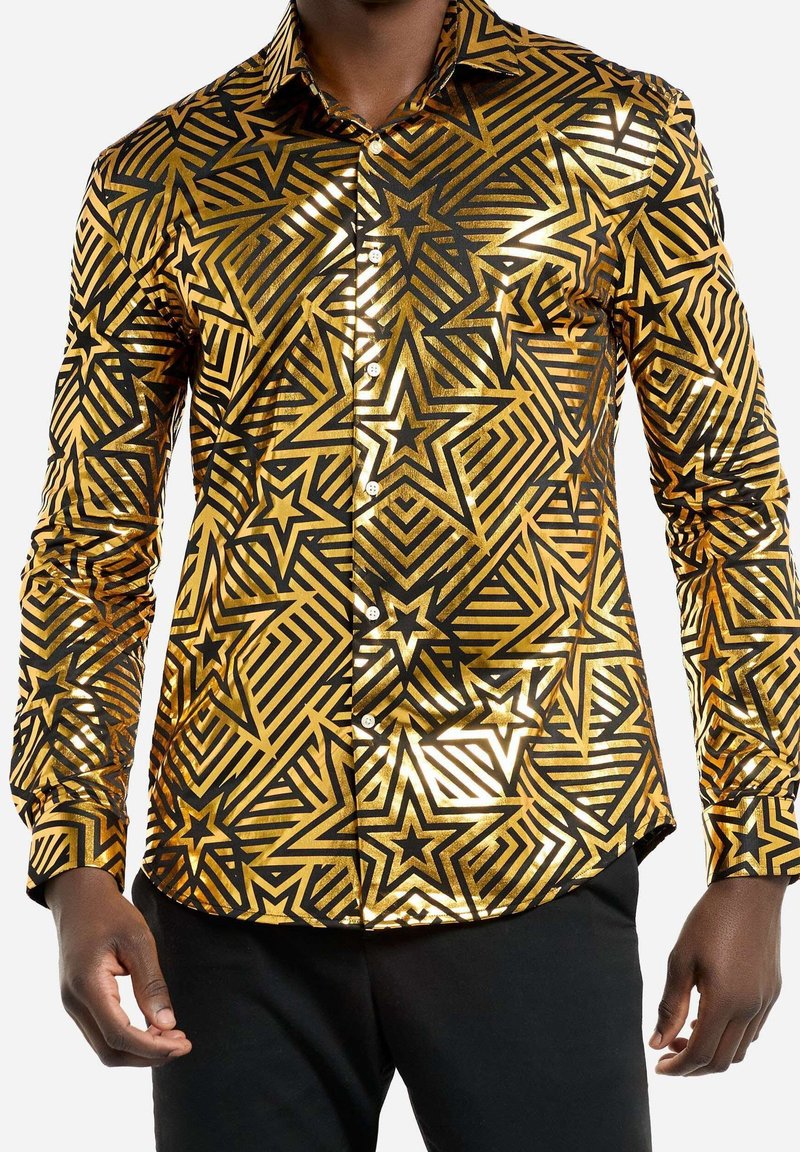 Chemise dorée brillante à manches longues avec des motifs géométriques d’étoiles noires portée avec un pantalon noir.