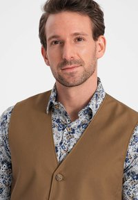 Hnědá látková vesta s výstřihem do V, opatřená jedním knoflíkem. Nosená s modrou košilí s květinovým vzorem pod ní.