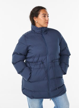 Kvinde iført en marineblå dunjakke med høj krave og snøre i taljen, stående med hænderne i lommerne, kigger til siden.