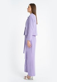 Liliowy dwuczęściowy garnitur z dopasowaną marynarką i szerokimi spodniami. Posiada strukturalny krój, gładką teksturę oraz szczegół z wiązaniem z boku.