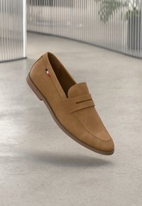 Tan suede loafers with a rounded toe, featuring a front strap and a small tricolor accent. Set against a neutral background.