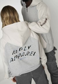 Mikiny s kapucí v světle šedé a krémové barvě, s vyšitým textem "M PLY APPAREL" a "SINCE 2016," s volným střihem a zipovým zapínáním.