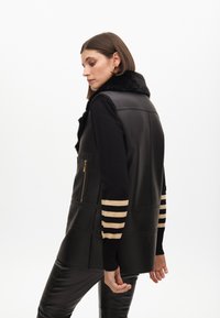 Chaleco de cuero negro con cuello de piel, que presenta un bolsillo con cremallera y mangas con rayas doradas. Textura suave con un ajuste moderno y a medida.