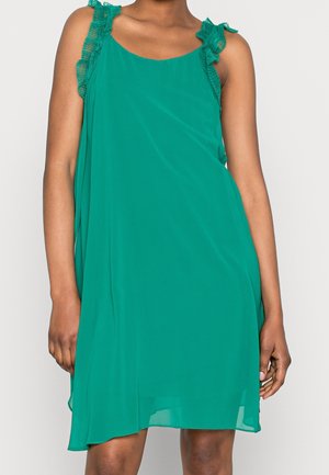 Femme portant une robe verte sans manches avec des bretelles volantées, debout devant un fond clair uni.