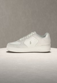 Λευκό sneaker χαμηλού ύψους με κορδόνια, πάνελ από σουέτ και δέρμα, και ένα μικρό κεντημένο λογότυπο στη πλευρά, που ακουμπά σε φωτεινή επιφάνεια.