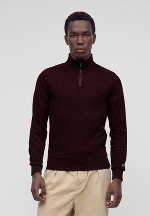 Homme portant un pull demi-fermeture éclair bordeaux foncé et un pantalon beige clair, debout devant un fond gris clair uni.