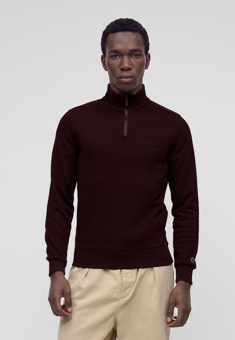 Homme portant un pull demi-fermeture éclair bordeaux foncé et un pantalon beige clair, debout devant un fond gris clair uni.