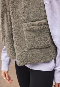 Vesta pufoasă de culoare verde măsliniu, dintr-un material fleece texturat, cu un buzunar frontal, purtată deasupra unei sweat-shirt de culoare gri deschis.
